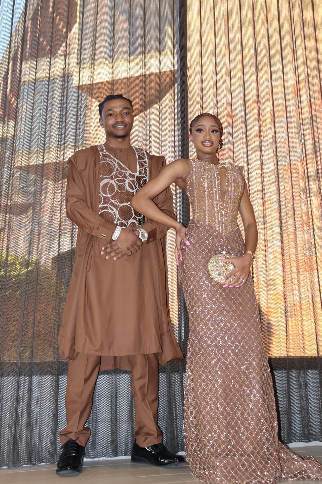 Couple in coordinated brown and gold formal wear in front of a shimmering curtain