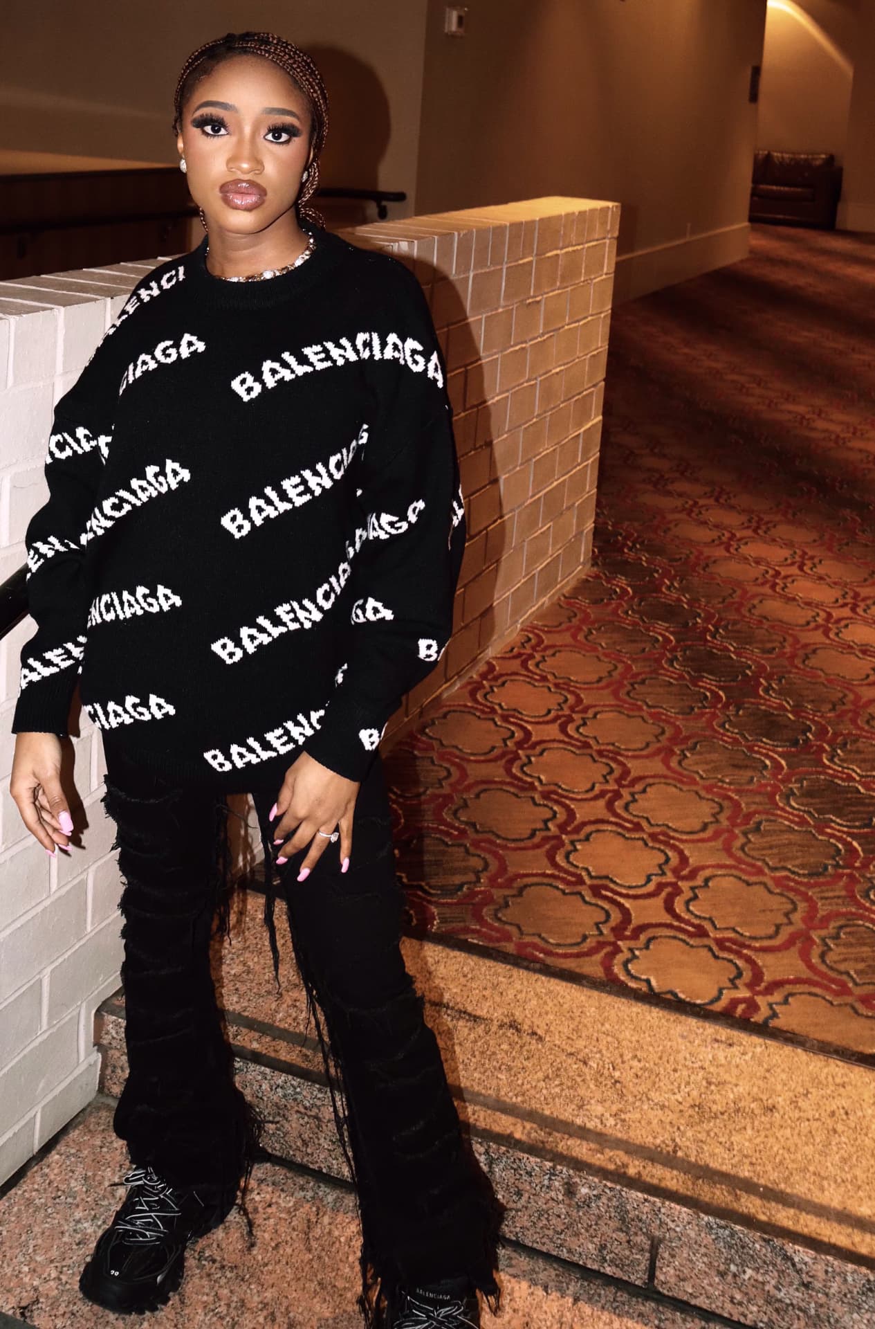 Full-length portrait in black designer knitwear against a brick wall